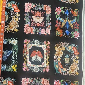 May include: A black fabric featuring a repeating pattern of rectangular frames containing various insects, including beetles and moths, surrounded by colorful floral arrangements. A yellow measuring tape showing 17 inches is visible on the left side.