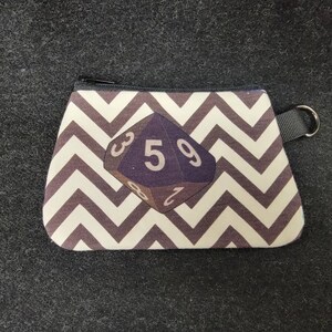 May include: A small zippered pouch with a black and white chevron pattern. The pouch features a purple die with the numbers 3, 5, and 9 visible.