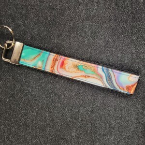 May include: A colorful key chain with a marbled design in shades of blue, pink, and gold. The key chain has a silver metal ring and a black fabric strap.