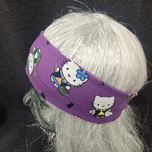 May include: A purple headband featuring a repeating pattern of a cartoon cat character. The headband is wrapped around a mannequin head with white hair. The cat character is wearing different outfits and has a blue flower on its ear.