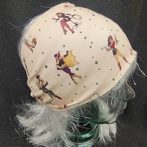 May include: A beige headband with a white elastic band. The headband has a repeating pattern of women in superhero costumes and stars.
