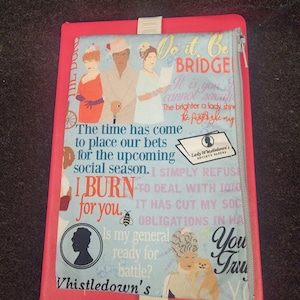 May include: A rectangular notebook with a bright pink cover and an elastic band, featuring a light blue fabric sleeve printed with illustrations of three figures, a small dog, a bee, and text including "I BURN for you" and "Lady Whistledown's Society Papers."