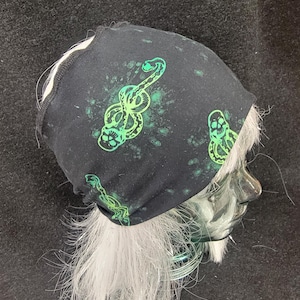 May include: Black beanie with a green snake and skull design. The beanie has a splatter effect and is worn on a mannequin head with white hair. The design features a stylized snake intertwined with a skull.