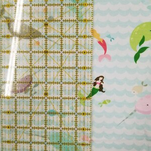 May include: A white fabric with a repeating pattern of blue waves, mermaids, narwhals, and fish. A ruler is placed on the fabric to show the scale.