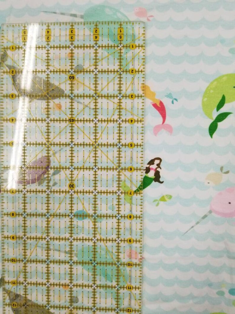May include: A white fabric with a repeating pattern of blue waves, mermaids, narwhals, and fish. A ruler is placed on the fabric to show the scale.