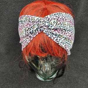May include: A white headband with a black and pink leopard print design. The headband is twisted and tied in a knot at the top.