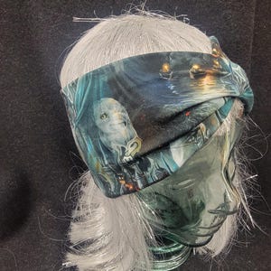 May include: A fabric headband with a fantasy-themed print featuring an owl and boats on a body of water. The headband is wrapped around a mannequin head with silver hair. The color palette includes shades of blue, green, and brown.