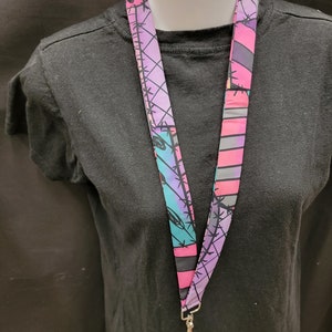 May include: A lanyard with a colorful pattern of barbed wire, pink, purple, and teal. The lanyard is hanging around a black t-shirt. The lanyard has a silver-colored metal clasp.