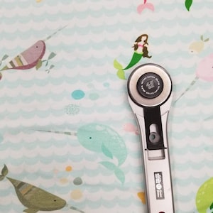 May include: A silver rotary cutter with a black handle and a 45 mm blade, on a light blue fabric with a pattern of narwhals, mermaids, and bubbles.