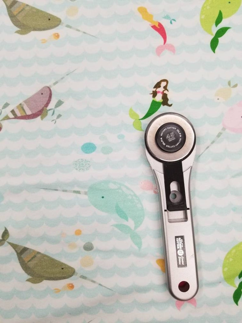 May include: A silver rotary cutter with a black handle and a 45 mm blade, on a light blue fabric with a pattern of narwhals, mermaids, and bubbles.