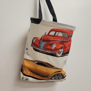 May include: Cream-colored tote bag featuring illustrations of classic cars in red and gold. The bag has black straps and a black trim at the top. The cars are detailed with chrome accents and white tires.