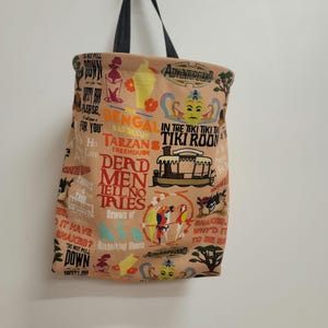 May include: A beige canvas tote bag with a black handle. The bag is printed with a variety of colorful images and text, including "Adventureland", "Bengal Barbecue", "Tarzan's Treehouse", "The Enchanted Tiki Room", "Derd Men Tell No Tries", and "Beware of Hitchhiking Ghosts".