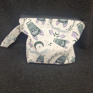May include: A white zippered pouch with a spooky bat print. The bats are purple and gray, with the words "Spooky 4 Life" and other Halloween-themed graphics. The pouch has a black zipper and a small handle.