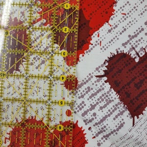 May include: White fabric with a red and gray splatter pattern. A ruler is placed on the fabric to show the scale of the pattern.