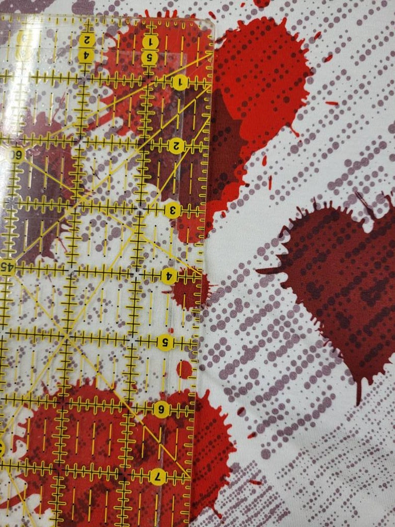 May include: White fabric with a red and gray splatter pattern. A ruler is placed on the fabric to show the scale of the pattern.