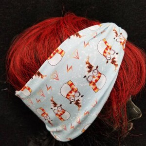 May include: A light blue headband with a repeating pattern of snowmen wearing glasses and scarves. The snowmen are wearing red and yellow scarves and have brown eyes. The background is a light blue with white snowflakes and lightning bolts.