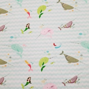 May include: A light blue fabric with a repeating pattern of narwhals, mermaids, and fish. The narwhals are green, pink, and brown. The mermaids are wearing pink and green dresses. The fish are red, yellow, and blue.