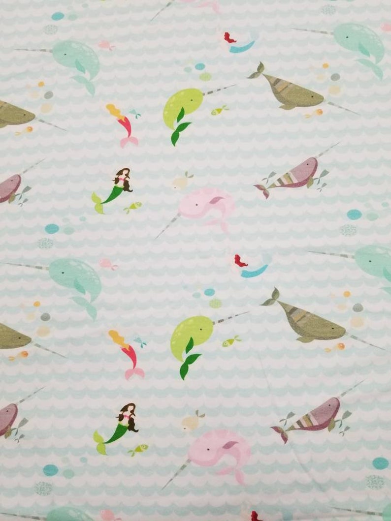 May include: A light blue fabric with a repeating pattern of narwhals, mermaids, and fish. The narwhals are green, pink, and brown. The mermaids are wearing pink and green dresses. The fish are red, yellow, and blue.