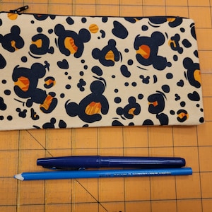 May include: A beige zippered pouch with a black and orange Mickey Mouse print. The pouch is lying on a brown gridded surface.