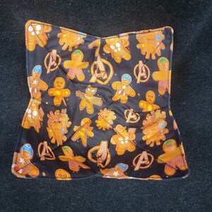 May include: Black quilted bowl cozy featuring a repeating pattern of orange gingerbread figures and the Avengers logo. The cozy is square with a folded design, and the edges are trimmed with a contrasting orange and white pattern.