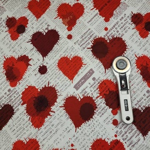 May include: Red heart-shaped splatter pattern fabric with a white background and a small silver rotary cutter on top.