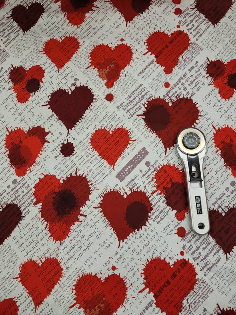 May include: Red heart-shaped splatter pattern fabric with a white background and a small silver rotary cutter on top.