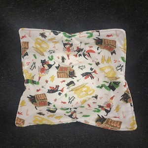 May include: A white, square, fabric bowl cozy with a whimsical pattern of black cats, red and gold treasure chests, and baking tools. The cozy is designed to hold a bowl of food and protect hands from heat.