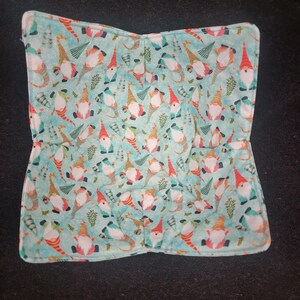 May include: A folded, square, light blue fabric item with a repeating pattern of gnomes wearing red hats and holding lanterns. The gnomes are set against a background of small green sprigs. The edges of the fabric are white.