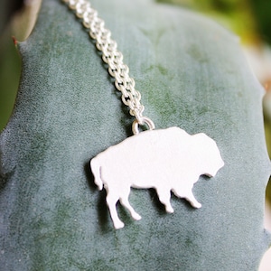 May include: A silver-toned necklace featuring a flat, solid pendant shaped like a buffalo in profile. The pendant is attached to a delicate, fine-link silver-toned chain that extends upward from the top of the animal figure's back.