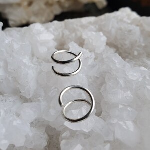 May include: Two silver nose rings, one is a simple hoop and the other is a double hoop, both are displayed on a white crystal background.