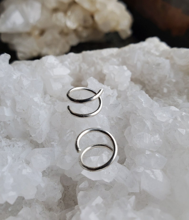 May include: Two silver nose rings, one is a simple hoop and the other is a double hoop, both are displayed on a white crystal background.