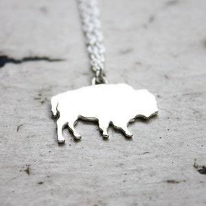 May include: A silver-toned necklace featuring a flat, solid pendant shaped like a bison in profile. The pendant is attached to a delicate, thin silver-toned chain that extends upward from the top of the bison's back, creating a simple, reflective metallic jewelry piece.