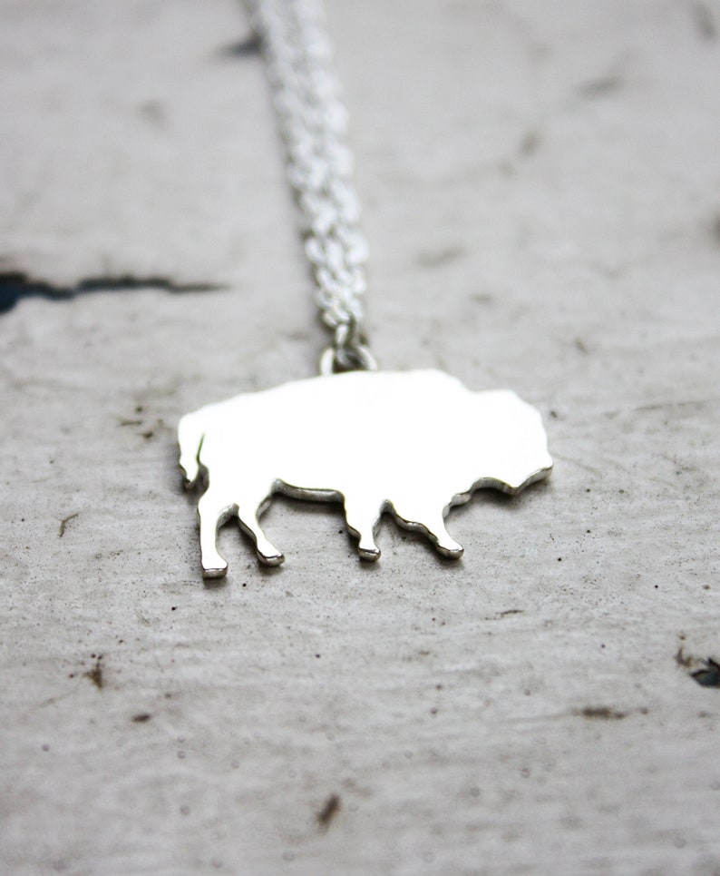 May include: A silver-toned necklace featuring a flat, solid pendant shaped like a bison in profile. The pendant is attached to a delicate, thin silver-toned chain that extends upward from the top of the bison's back, creating a simple, reflective metallic jewelry piece.
