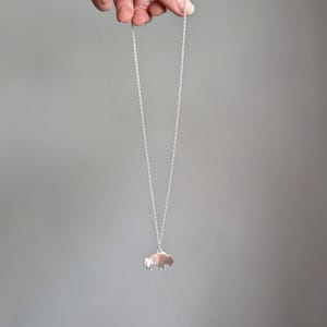 May include: A silver-toned necklace featuring a small, flat pendant shaped like a bison in profile. The pendant hangs from a delicate, thin silver-colored metal chain with small circular links. The design is simple, metallic, and reflective.