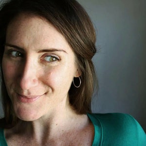 May include: A woman with brown hair wearing a green v-neck shirt and gold hoop earrings. She is looking to the side and smiling.