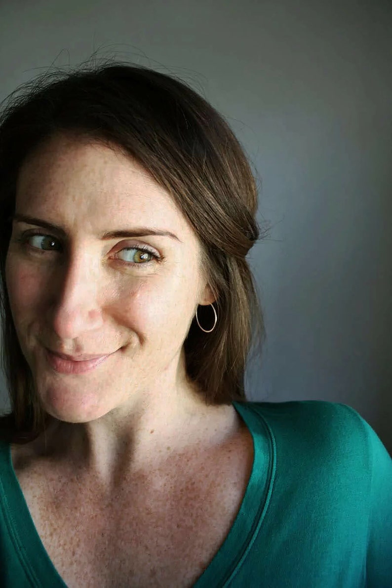 May include: A woman with brown hair wearing a green v-neck shirt and gold hoop earrings. She is looking to the side and smiling.