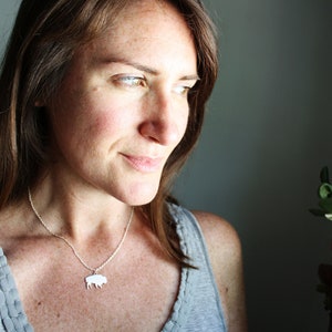 May include: A silver-toned necklace featuring a small, flat, silhouette-style pendant shaped like a bison. The pendant hangs from a delicate, thin metal chain that rests against the collarbone, providing a simple and clean aesthetic.