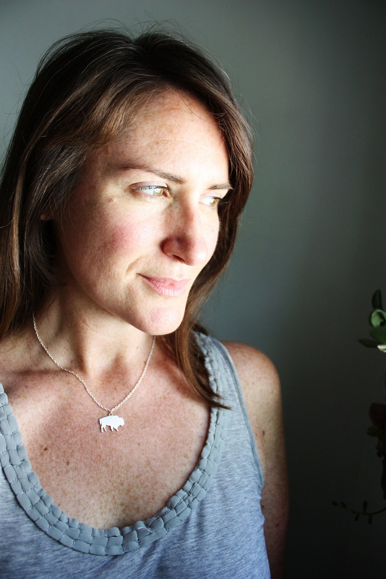 May include: A silver-toned necklace featuring a small, flat, silhouette-style pendant shaped like a bison. The pendant hangs from a delicate, thin metal chain that rests against the collarbone, providing a simple and clean aesthetic.