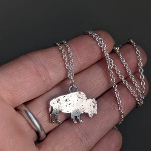 May include: A silver-toned necklace featuring a pendant shaped like a bison. The pendant has a textured, speckled surface finish and is attached to a delicate silver-toned metal chain with small, round, interlocking links.