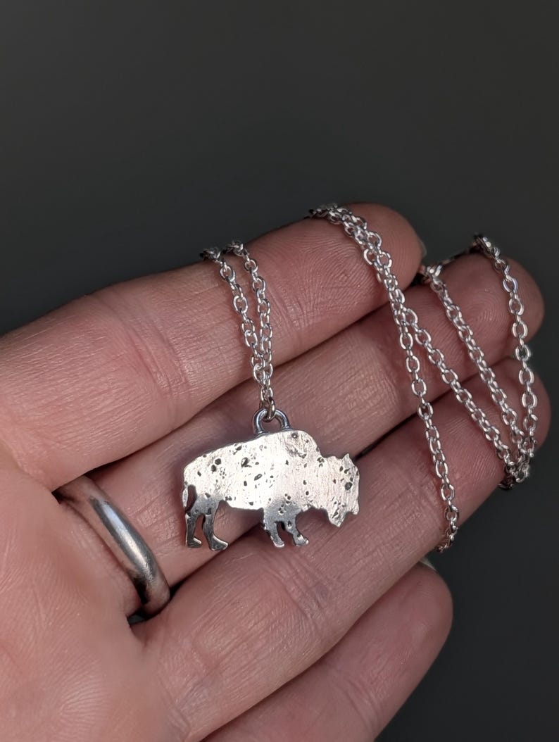 May include: A silver-toned necklace featuring a pendant shaped like a bison. The pendant has a textured, speckled surface finish and is attached to a delicate silver-toned metal chain with small, round, interlocking links.