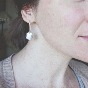 May include: A close-up of a person wearing a silver hammered disc earring. The earring is hanging from the person's earlobe and is a simple, minimalist design.