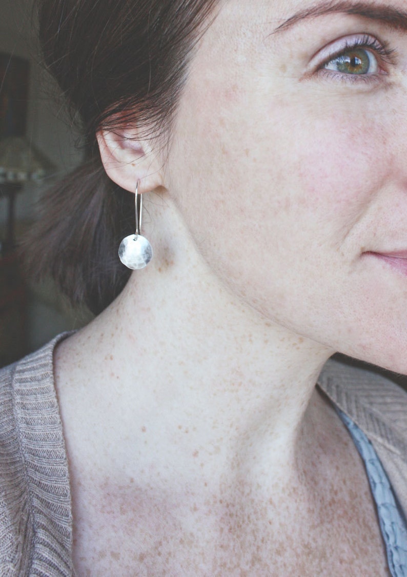 May include: A close-up of a person wearing a silver hammered disc earring. The earring is hanging from the person's earlobe and is a simple, minimalist design.