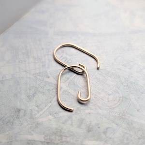 May include: A pair of rose gold-toned metal earrings, each featuring an elongated, open oval shape with a curved hook end. The earrings have a smooth, polished finish and a minimalist, continuous wire design that forms a rounded rectangular loop.