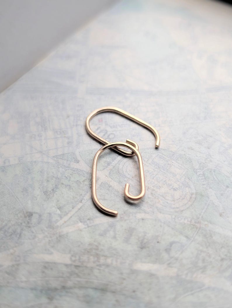 May include: A pair of rose gold-toned metal earrings, each featuring an elongated, open oval shape with a curved hook end. The earrings have a smooth, polished finish and a minimalist, continuous wire design that forms a rounded rectangular loop.