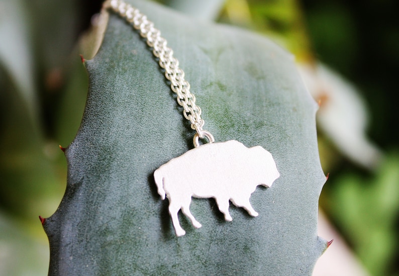 May include: A silver-toned necklace featuring a flat, solid pendant shaped like a buffalo in profile. The pendant is attached to a delicate, fine-link silver-toned chain that extends upward from the top of the animal figure's back.