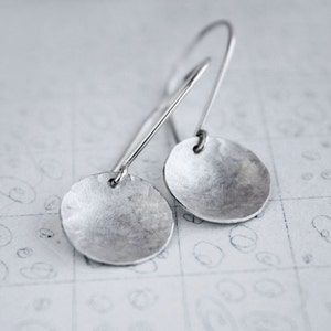 May include: A pair of silver dangle earrings with a hammered, circular design. The earrings have a simple, minimalist style.