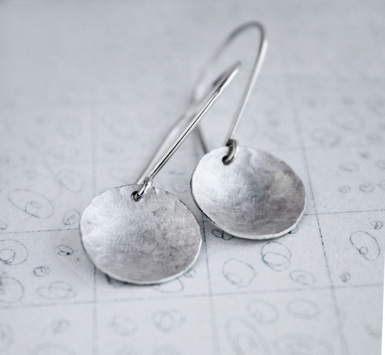 May include: A pair of silver dangle earrings with a hammered, circular design. The earrings have a simple, minimalist style.