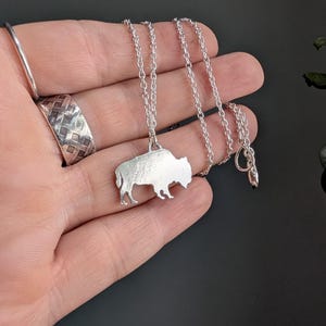May include: A silver-toned necklace featuring a pendant shaped like a buffalo in profile. The pendant hangs from a delicate silver-toned chain with small circular links. A wide silver-toned ring with a textured, woven pattern is also visible.