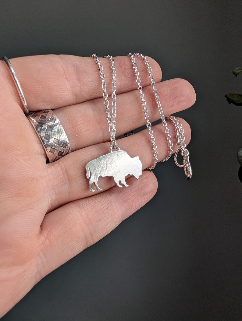May include: A silver-toned necklace featuring a pendant shaped like a buffalo in profile. The pendant hangs from a delicate silver-toned chain with small circular links. A wide silver-toned ring with a textured, woven pattern is also visible.