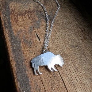 May include: A silver-toned metal pendant shaped like a buffalo silhouette, suspended from a matching silver-toned link chain. The pendant features a smooth, reflective surface and is attached to the chain by a small circular jump ring.